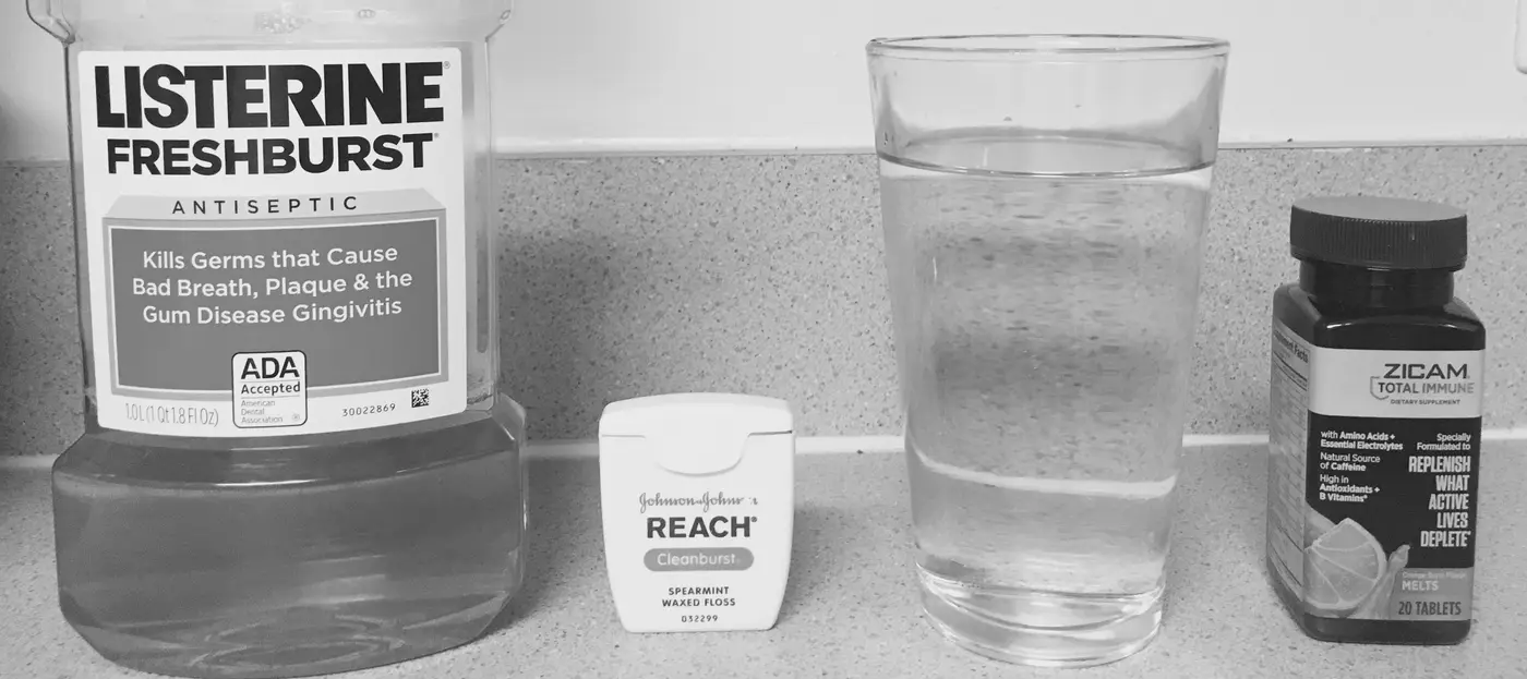 Black and white photo of ordinary bathroom items lined up on a counter: mouthwash, dental floss, a glass of water, and a supplement bottle.