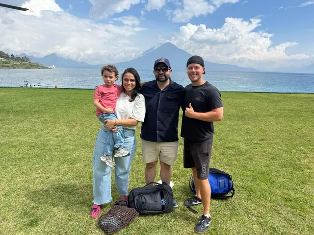 Zach standing on a lawn with Fernando, Lore, and her son, Lake Atitlán and a volcano rising behind them under a partly cloudy sky.