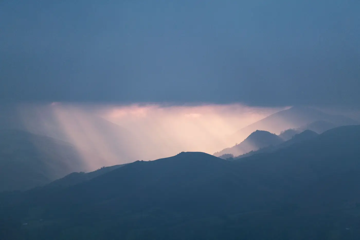 Muted light breaking through rain clouds over a range of mountains.