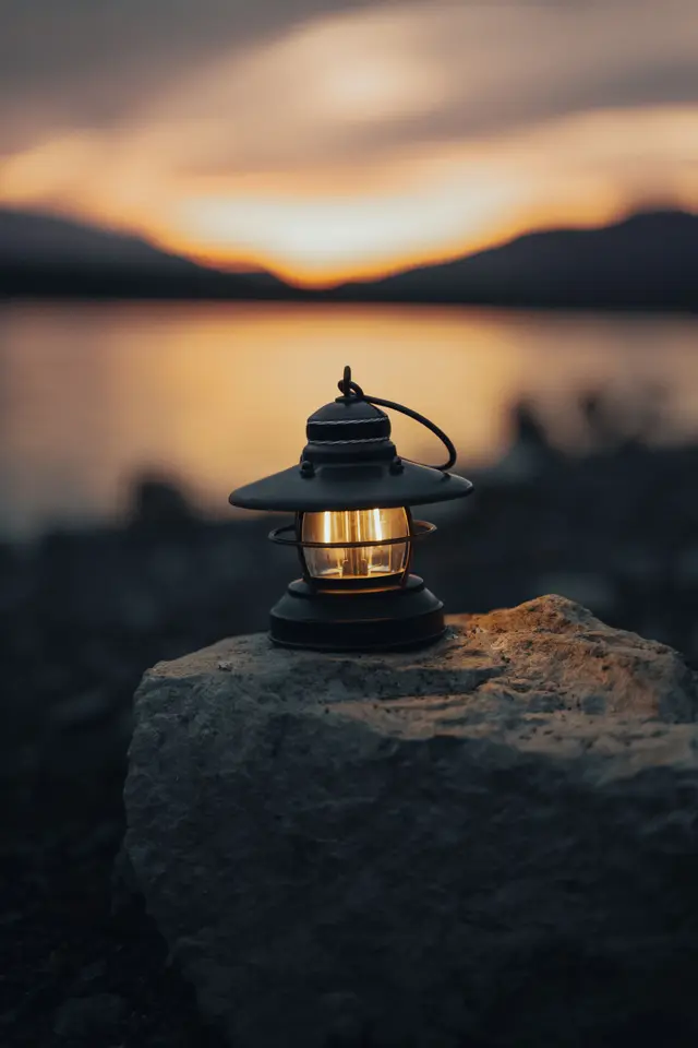 A small lit lantern glowing on a rock beside a lake at sunset.