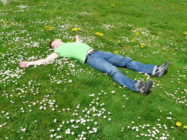 A man lying on his back in a grassy field full of daisies and dandelions, arms outstretched.