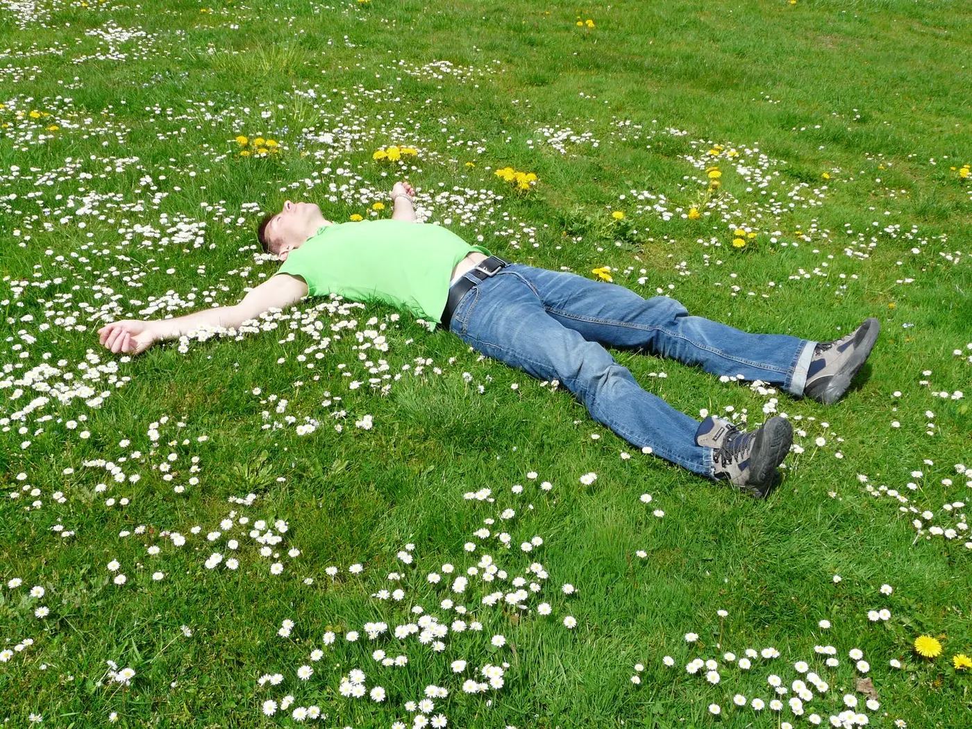 A man lying on his back in a grassy field full of daisies and dandelions, arms outstretched.