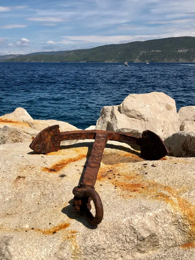 A weathered iron anchor resting on sunlit coastal rocks above the sea.