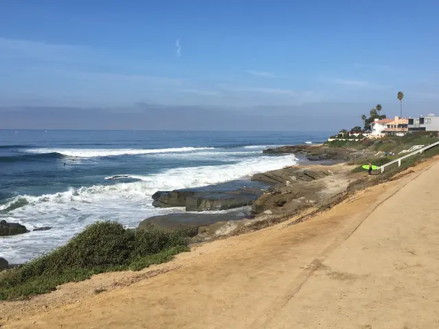 Waves breaking over rocky coastline below cliffside houses, with a person holding a surfboard on the path.