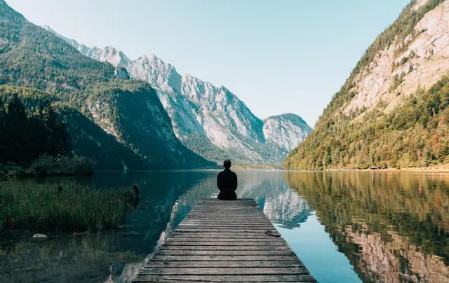 A person sitting at the end of a wooden dock on a still mountain lake, framed by tall peaks.