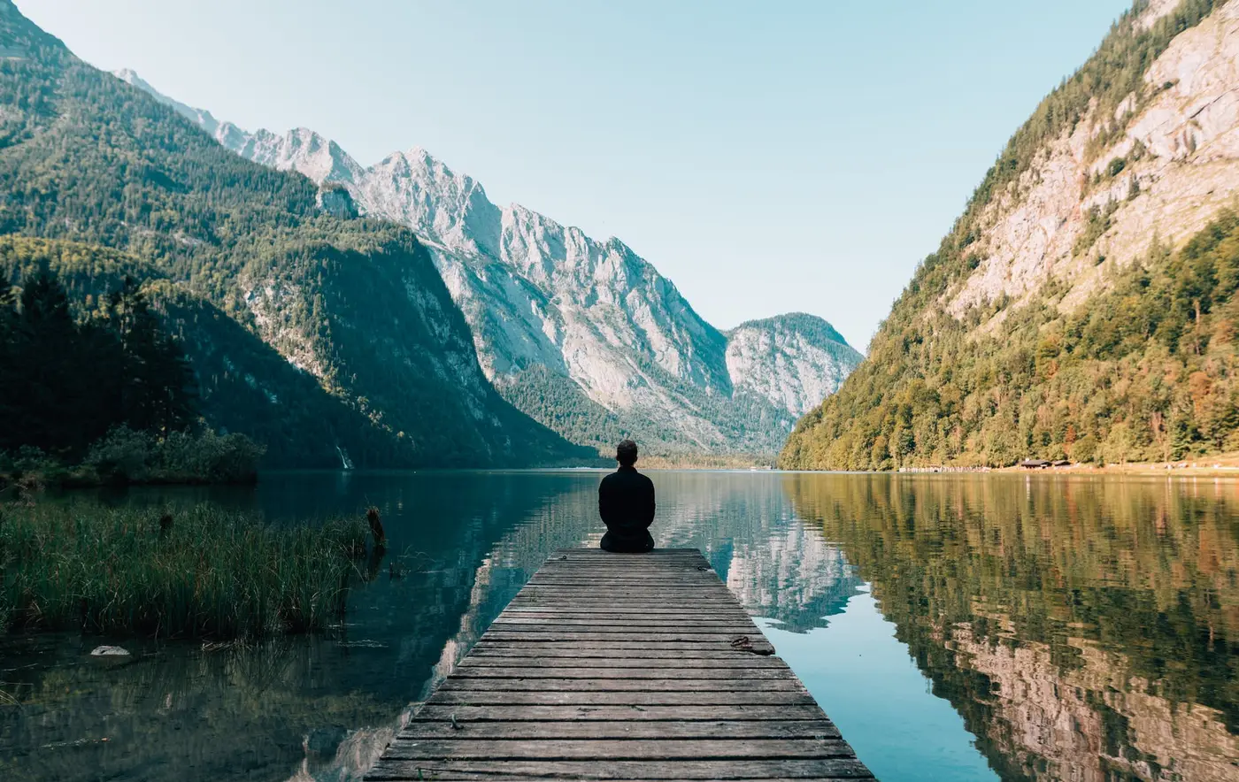 A person sitting at the end of a wooden dock on a still mountain lake, framed by tall peaks.