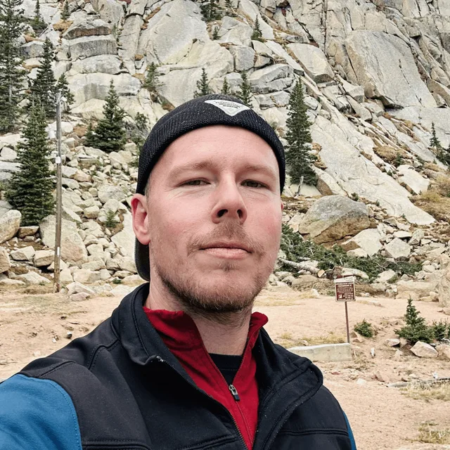 Zach Heern smiling outdoors in front of a rocky Colorado mountainside, wearing a beanie and a fleece vest.